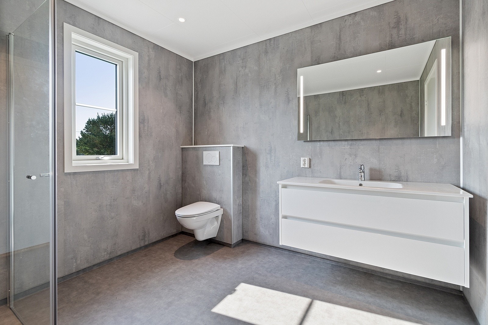 Moderne bad/vaskerom på 7,5 m² med vegghengt toalett, romslig servant med underskuffer, speil med belysning og dusj med glassdører. Galleribilde