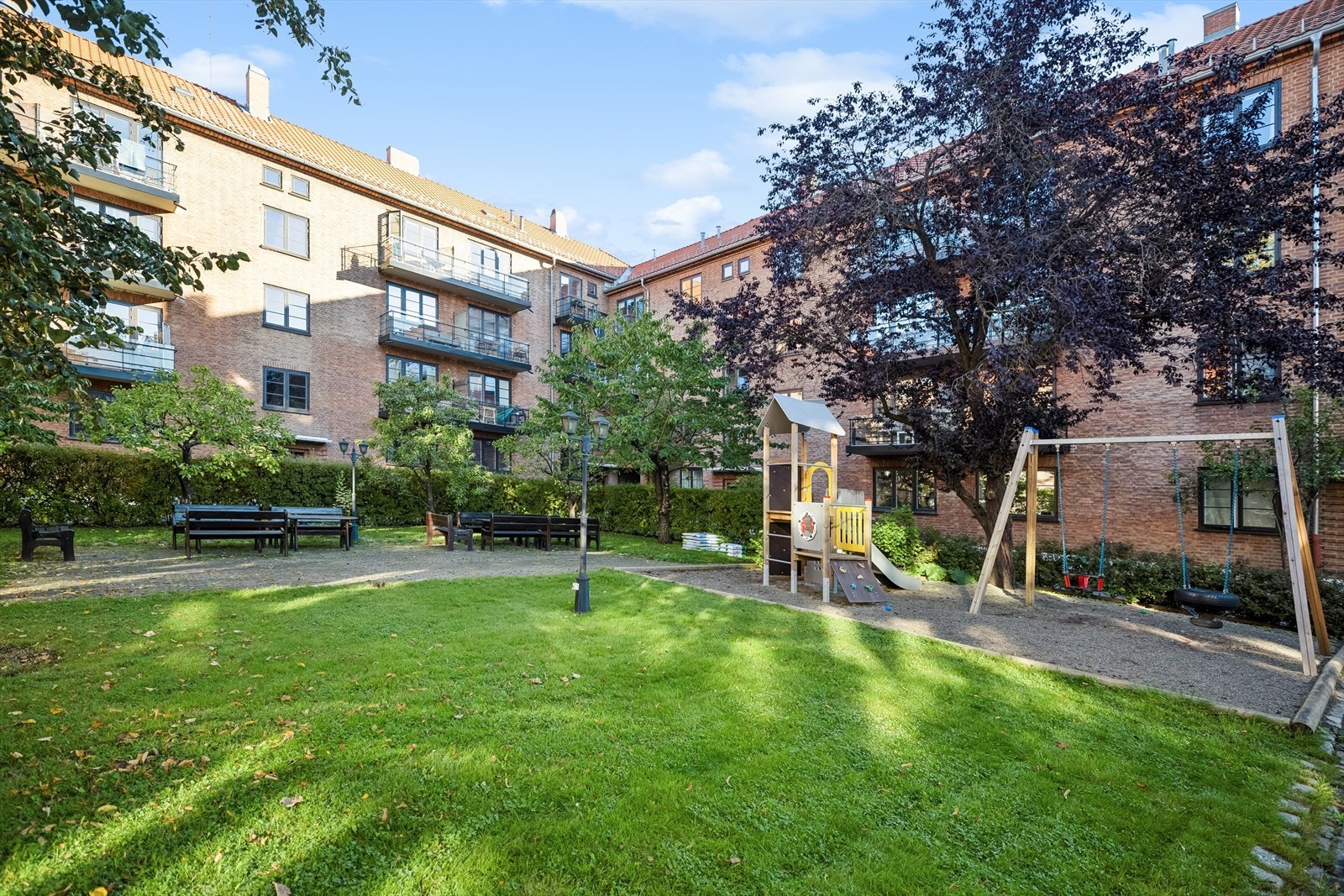 Bakgården er et hyggelig og velholdt fellesområde som inviterer til både avslapning og sosialt samvær - her finner du grønne planter, frukttrær og blomsterbed i tillegg til flere sittegrupper, felles grill og lekeplass med ulike apparater Galleribilde