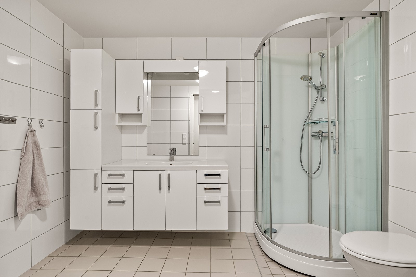 Badet har flislagte overflater på både gulv og vegger. Rommet er innredet med hvit baderomsinnredning som byr på god oppbevaringsplass i skuffer og skap. Over servanten er det montert speil som gir et lyst og funksjonelt preg. Galleribilde