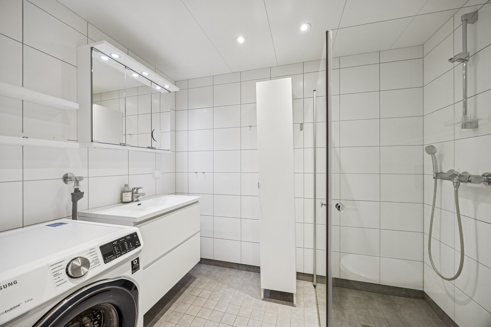 Flislagt bad med vegg montert toalett, dusj hjørne og opplegg for vaskemaskin. Vaskemaskin medfølger. Galleribilde