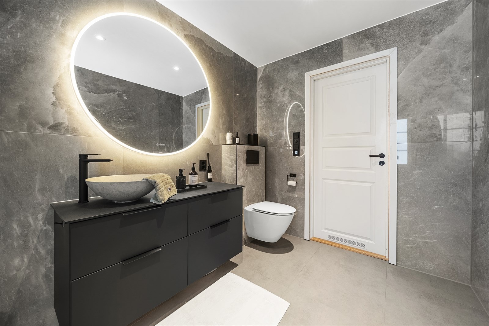 Badet er helfliset med tidsriktige store fliser og varmekabler i gulv. Badet er utstyrt med servant over skuffeinnredning, speil med belysning, vegghengt toalett og dusjhjørne med innfellbare glassvegger Galleribilde