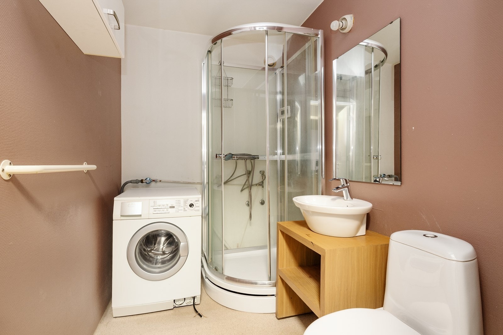 Bad fra byggetiden utstyrt med gulvmontert toalett, dusjkabinett og overliggende servant på innredning. Speil og enkel belysning på vegg. Galleribilde