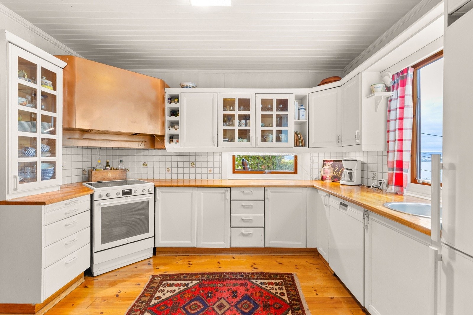 Kjøkkenet har en klassisk innredning med hvite profilerte fronter, laminert benkeplate med nedfelt vask, innebygde vitrineskap og vindu som gir rikelig arbeidslys. Galleribilde