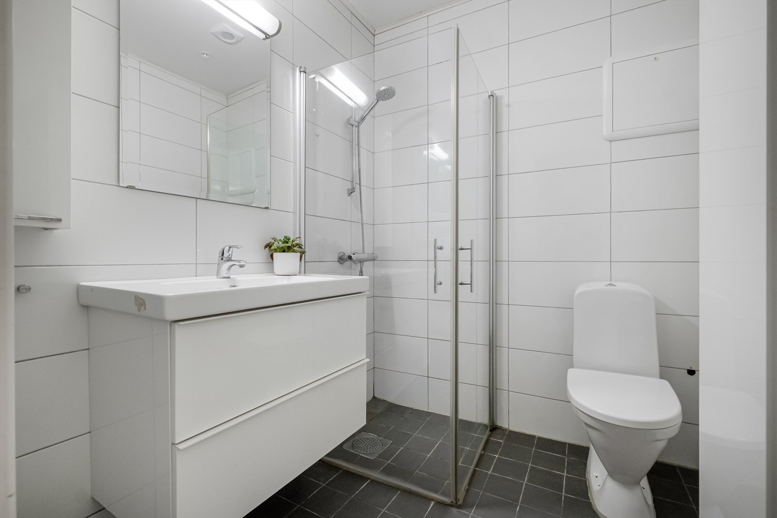 Flislagt baderom med servant, speil, servantinnredning med skuffer, dusjhjørne og toalett. Galleribilde