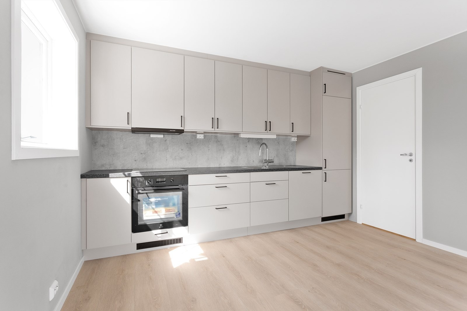 Kjøkkeninnredningen fra Epoq byr på god skap- og skuffeplass langs stueveggen, med glatte grå fronter og en mørk laminatbenkeplate som skaper en lekker kontrast. Galleribilde