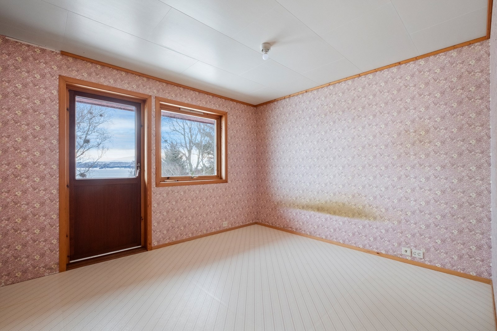 Tapetserte vegger og himlingsplater preger rommet, med synlige fuktskader på veggen. Utgang til balkong gir enkel tilgang til uteområdet. Galleribilde