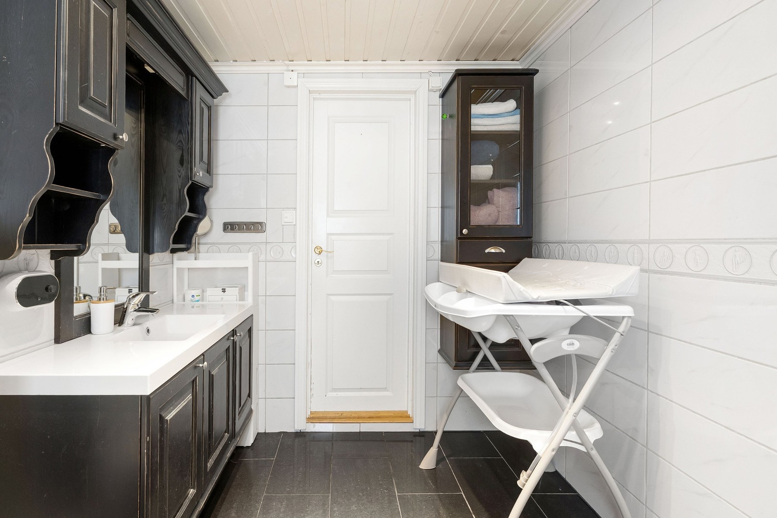 Bad med hvite fliser, sorte skap og servant. Lys takpanel og dør med messinghåndtak. Galleribilde