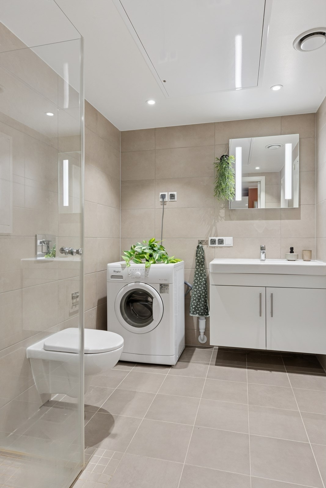 Moderne, flislagt bad med vegghengt toalett, dusjhjørne og opplegg for vaskemaskin. Galleribilde