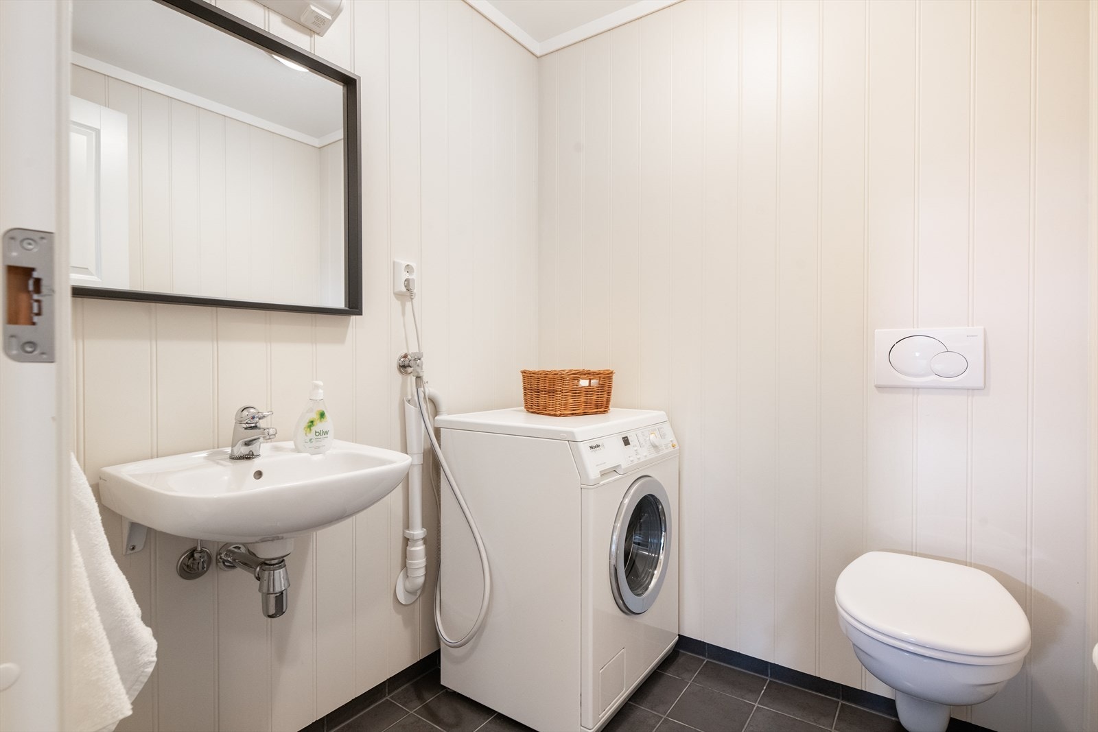 Toalettrom/vaskerom med flis på gulv og varmekabler. Rommet innehar servant, veggmontert toalett og opplegg for vaskemaskin. Galleribilde