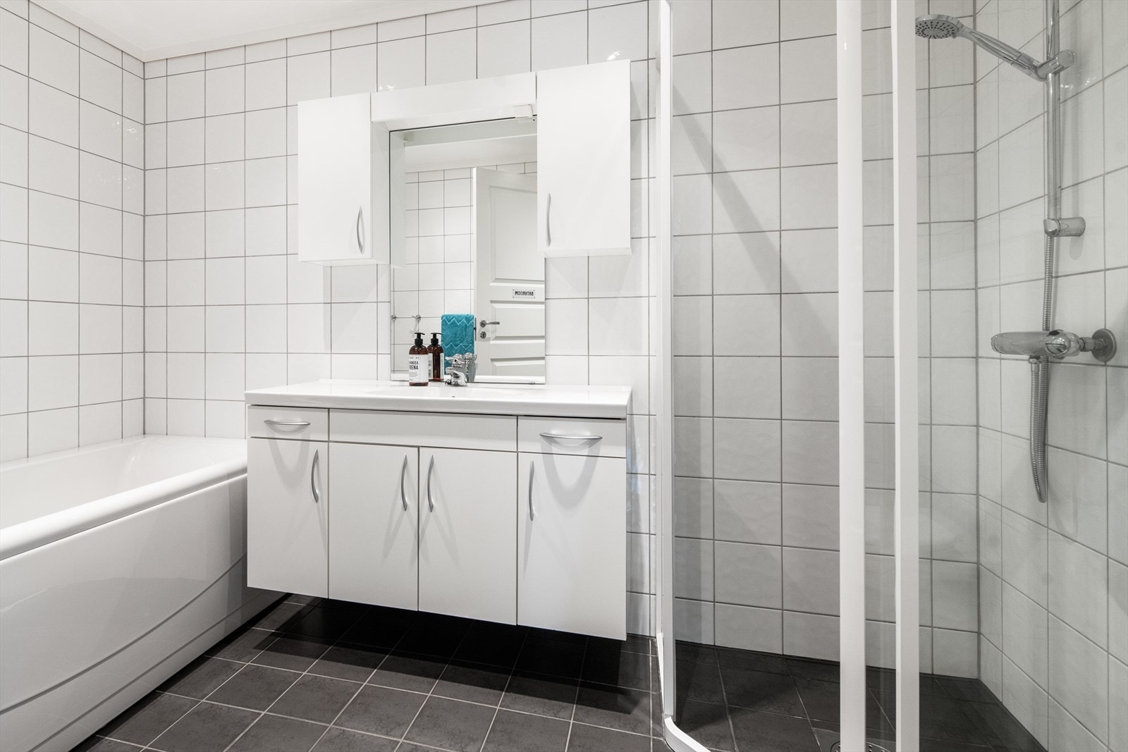 Flislagt bad med varmekabler i gulv. Badet er innredet med dusjhjørne, badekar og servantinnredning med skuffer, skap og speil. Galleribilde