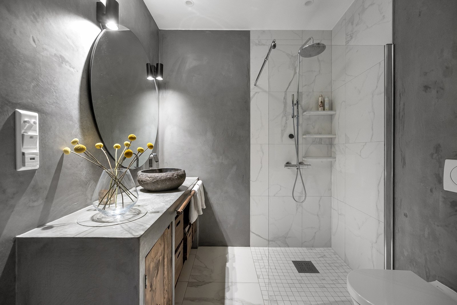 Badet er lekkert med mikrosementerte vegger og flisene fortsetter som en rød tråd fra resten av boligen. Dette gjentas i servantinnredningens benkeplate. Treskuffene er en fin kontrast til steinvask, sement og marmorflisene. Galleribilde