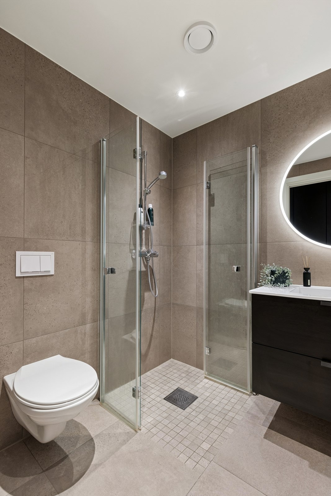 Moderne baderomsinnredning med sorte, glatte fronter og heldekkende servantplate. Over innredningen er det et rundt speil med integrert belysning. Galleribilde