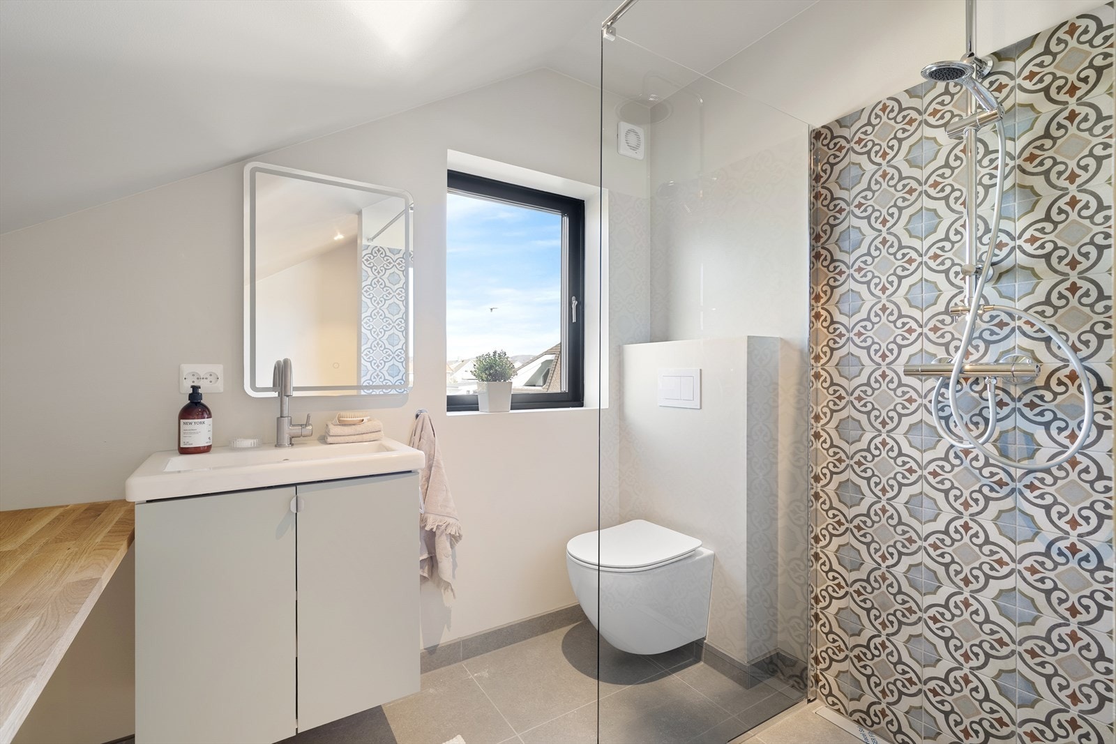 Badet med flislagt dusjhjørne med glassvegg, vegghengt toalett og servantskap med overliggende servant. Vegghengt lyssatt speil Galleribilde