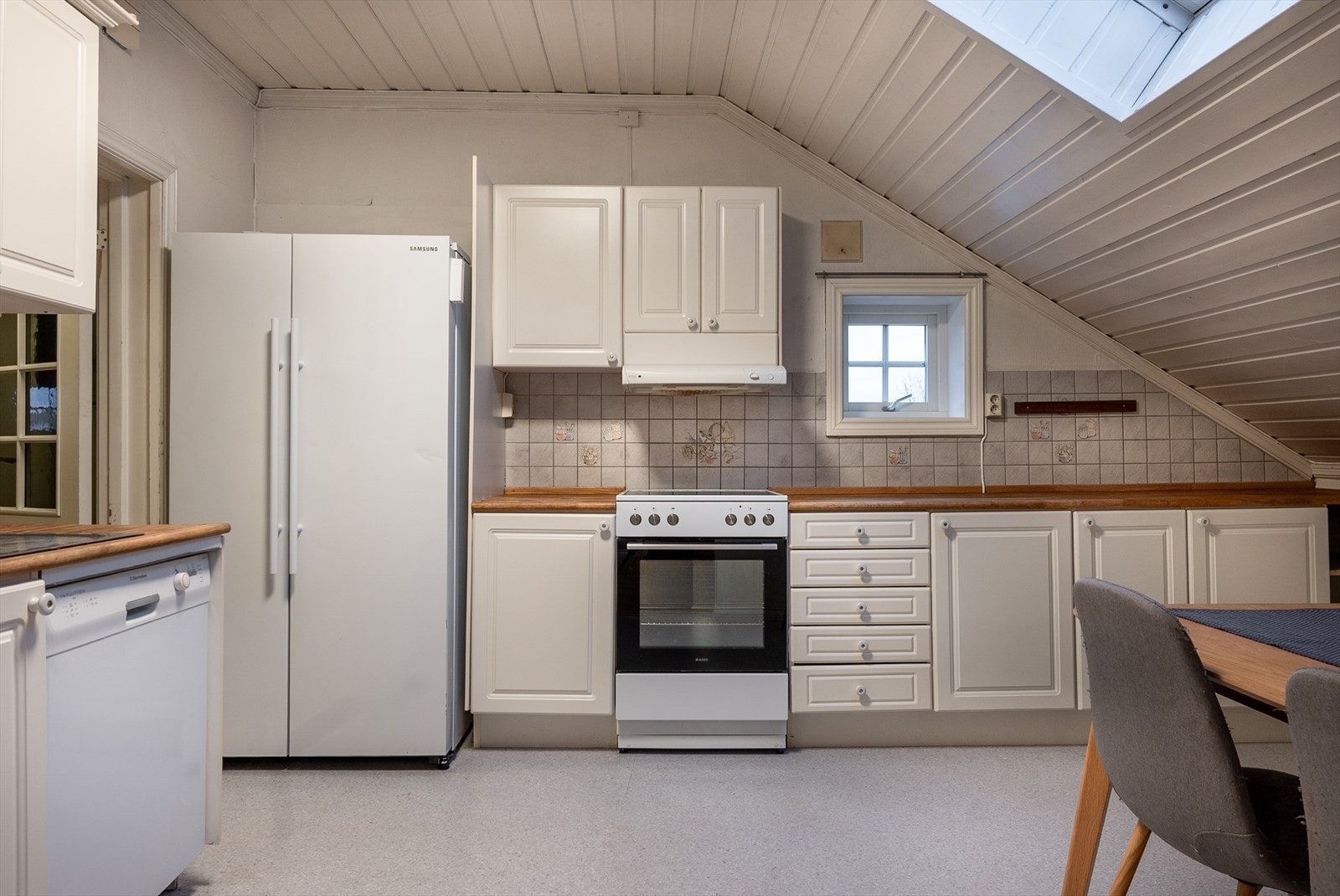 Kjøkkeninnredningen fyller to vegger _ med benkeplater i heltre, hvite fronter og frittstående hvitevarer. Her er et mindre veggvindu, men hoveddelen av dagslyset strømmer inn fra takvinduet. Galleribilde