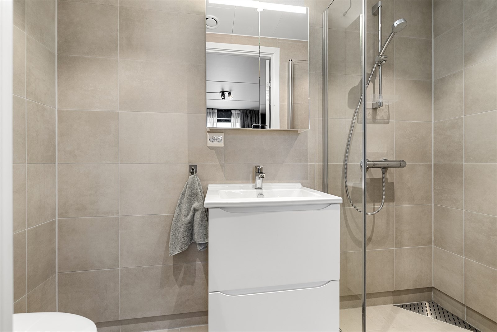 Badet er moderne og delikat med lyse fliser på gulv og vegger, dusjhjørne, vegghengt toalett og baderomsmøbel med tilhørende speilskap. Galleribilde