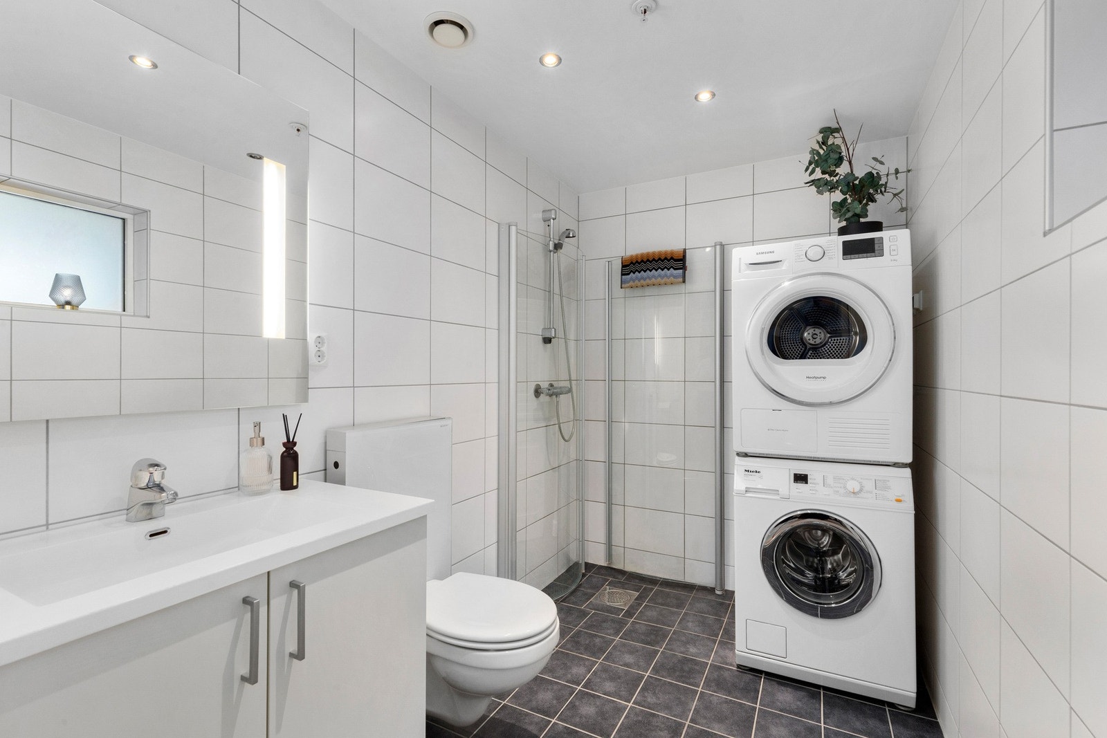 Badet er på 4,7 kvm og har innredning med dusjdører i glass, veggmontert WC, kran og avløp for vaskemaskin, og servantskap med gode oppbevaringsmuligheter. Galleribilde