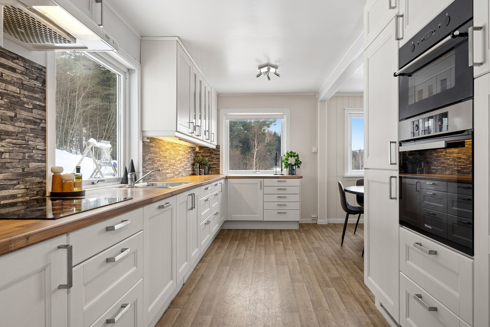 Innredningen har hvitlaserte, profilerte fronter på over- og underskap. Overskapene er takhøye for et helhetlig uttrykk og ekstra lagringsplass, og to av dem har glassfelt som gir kjøkkenet et lett og elegant preg. Galleribilde