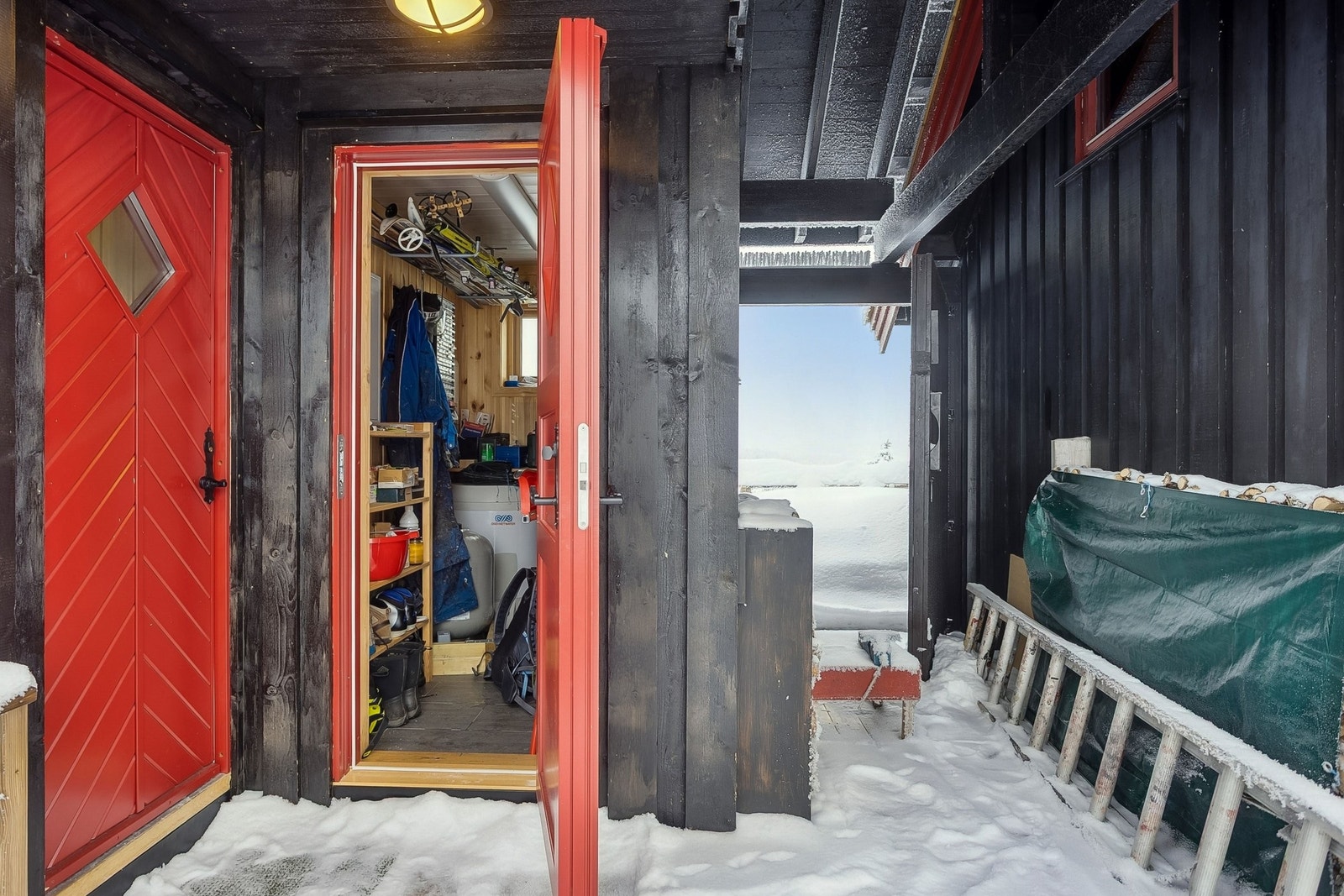 Åpen dør inn til teknisk rom som også har plass til vaskemaskin og smørebord. Svalgang mellom hovedhytta og tilbygget. Galleribilde