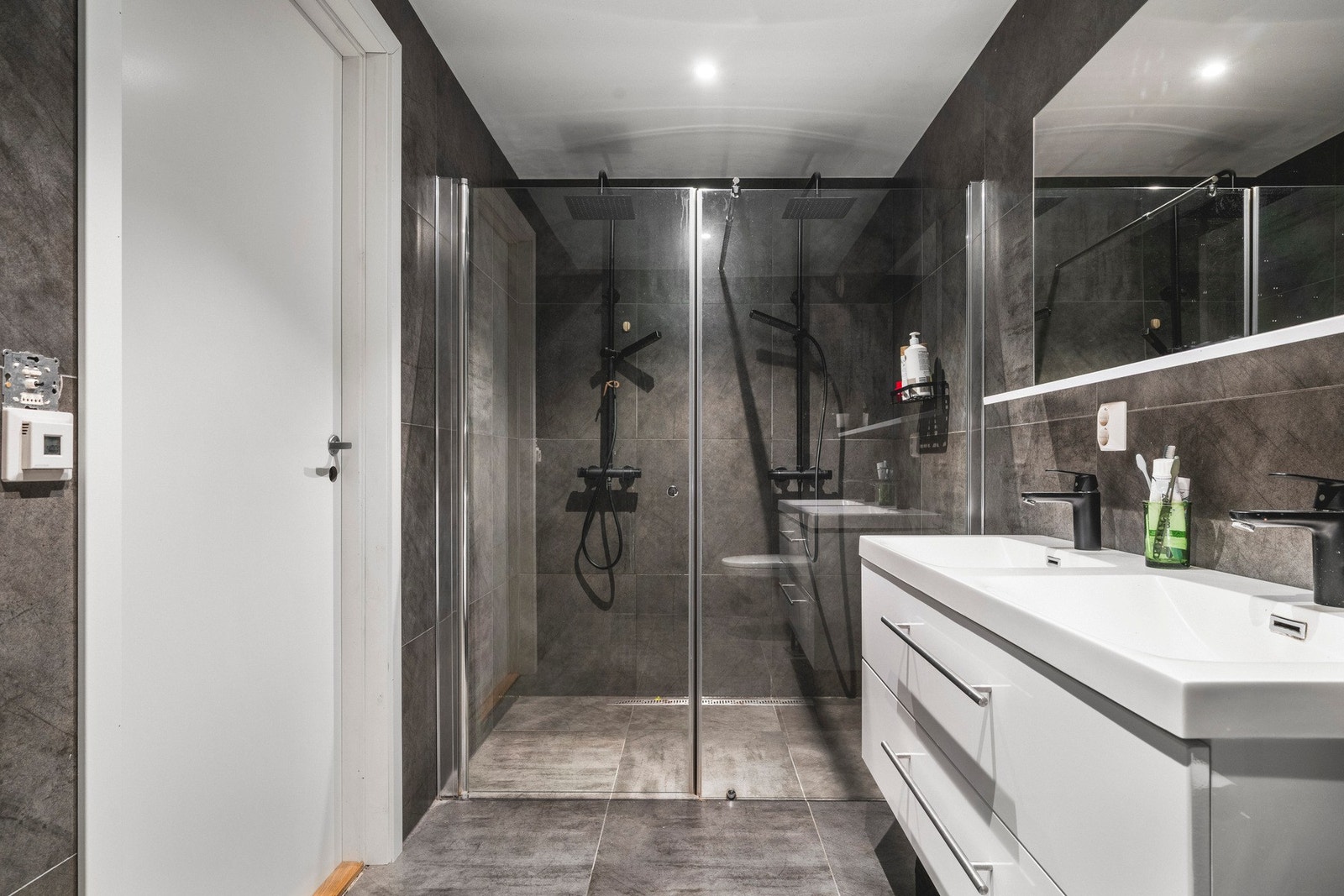 Badet inneholder vegghengt toalett, dobbel helstøpt servant, skuffer under servant med slette fronter og dusjdører i klart glass. Galleribilde