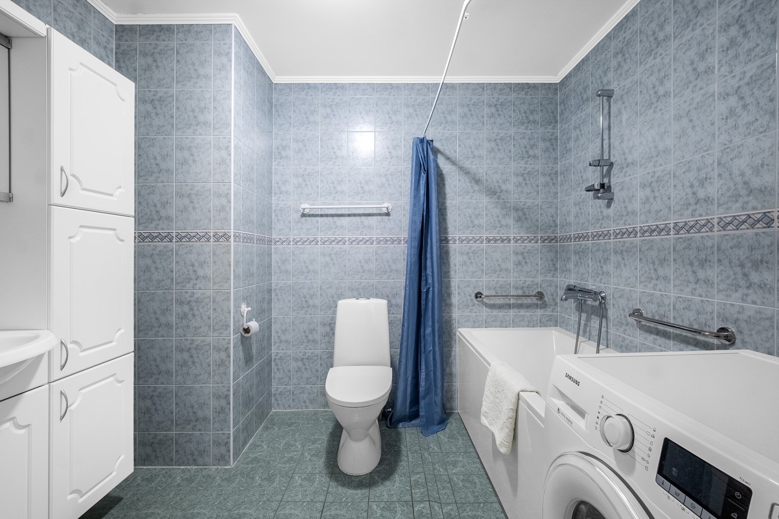 Videre fører gangen deg inn til et kombinert bade- og vaskerom. Badet er opprinnelig fra byggeåret og har fliser på både gulv og vegger, samt malt himling. Komforten ivaretas med elektriske varmekabler i gulvet. Galleribilde