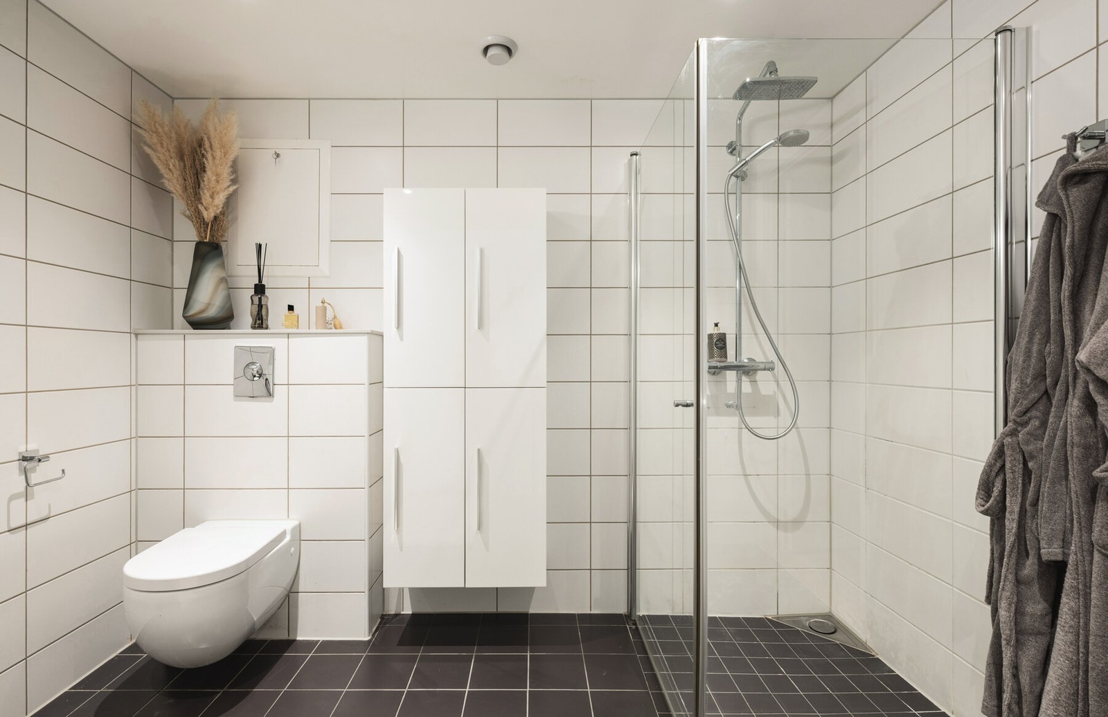 Bad med dusjhjørne, vegghengt toalett og skap. Flislagte vegger og gulv. Galleribilde