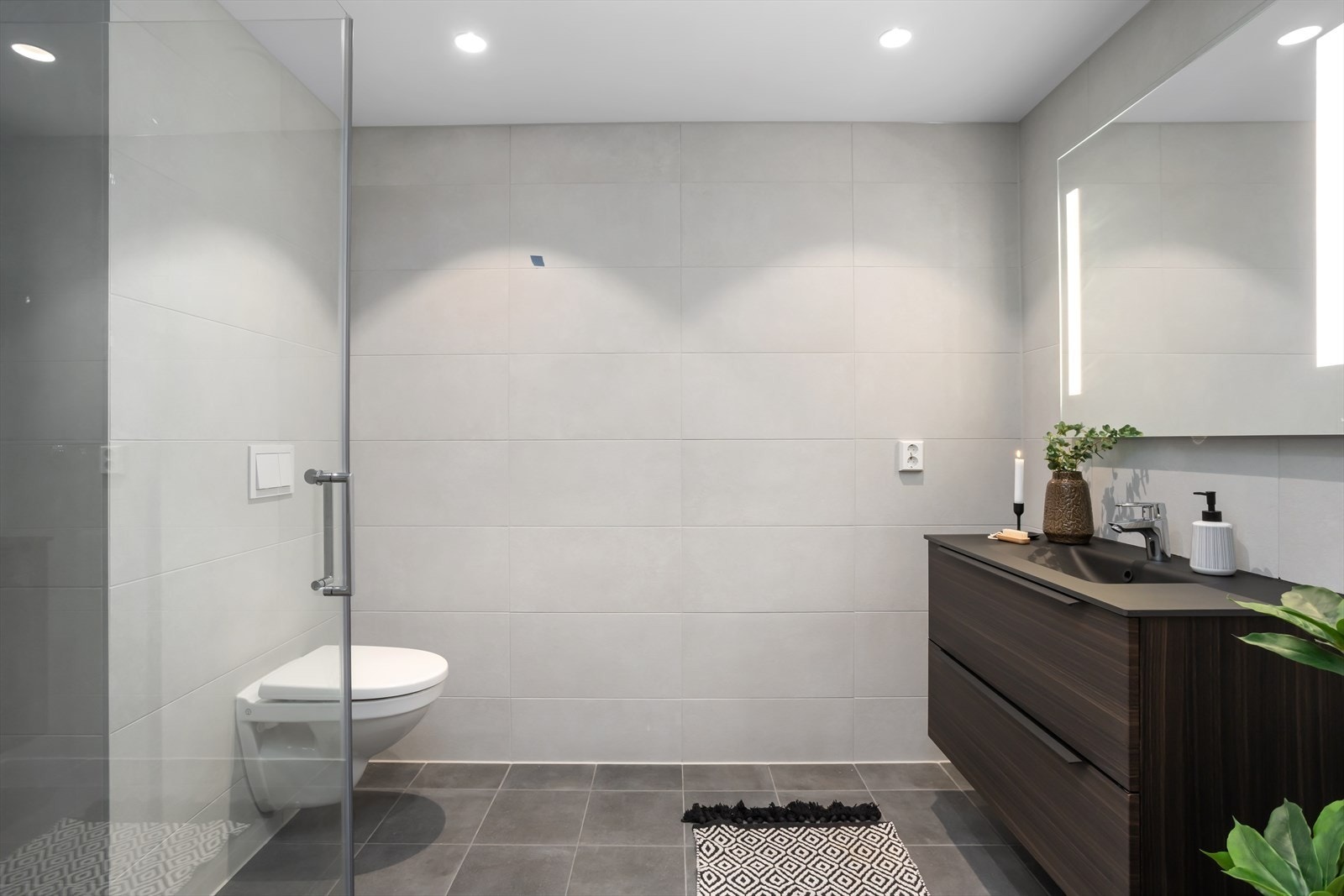 Baderommet tilknyttet entréen er pent flislagt med behagelig gulvvarme og downlights innfelt i himling. Baderommet er innredet med vegghengt toalett, mørk servantseksjon med dobbel skuff, speil med integrert belysning. Galleribilde