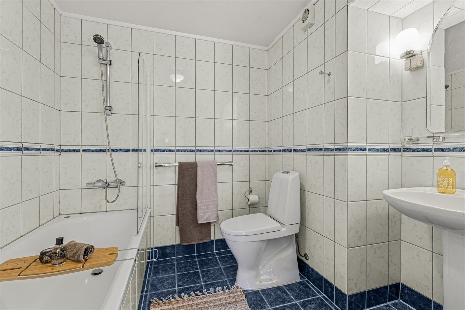 Baderom har fliser på gulv og vegger, inneholder: innfliset badekar, stående toalett og servant. Galleribilde