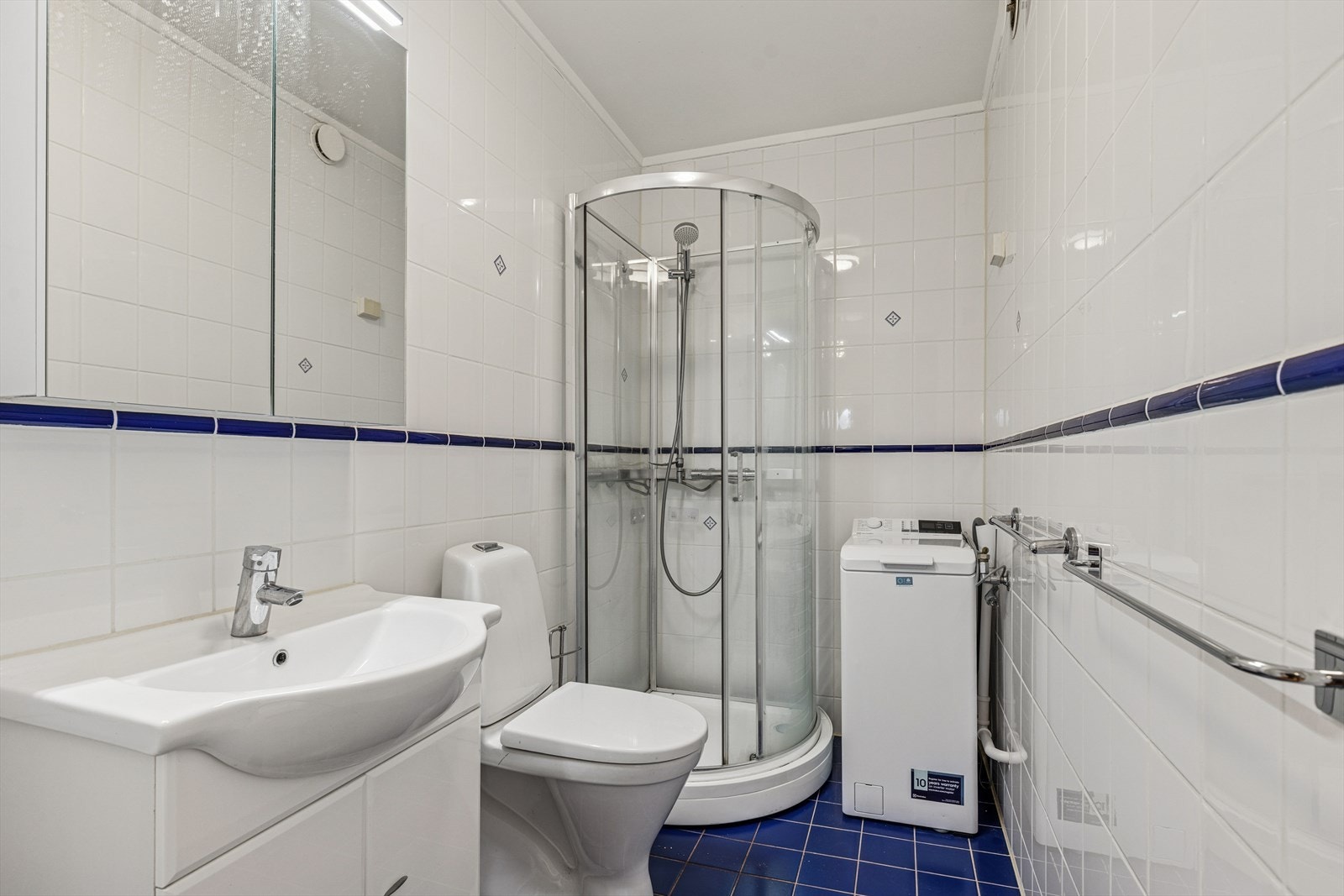 Baderom med fliser på gulv og vegger, med servant, stående toalett, dusj kabinett, og en areal effektiv vaskemaskin. Galleribilde