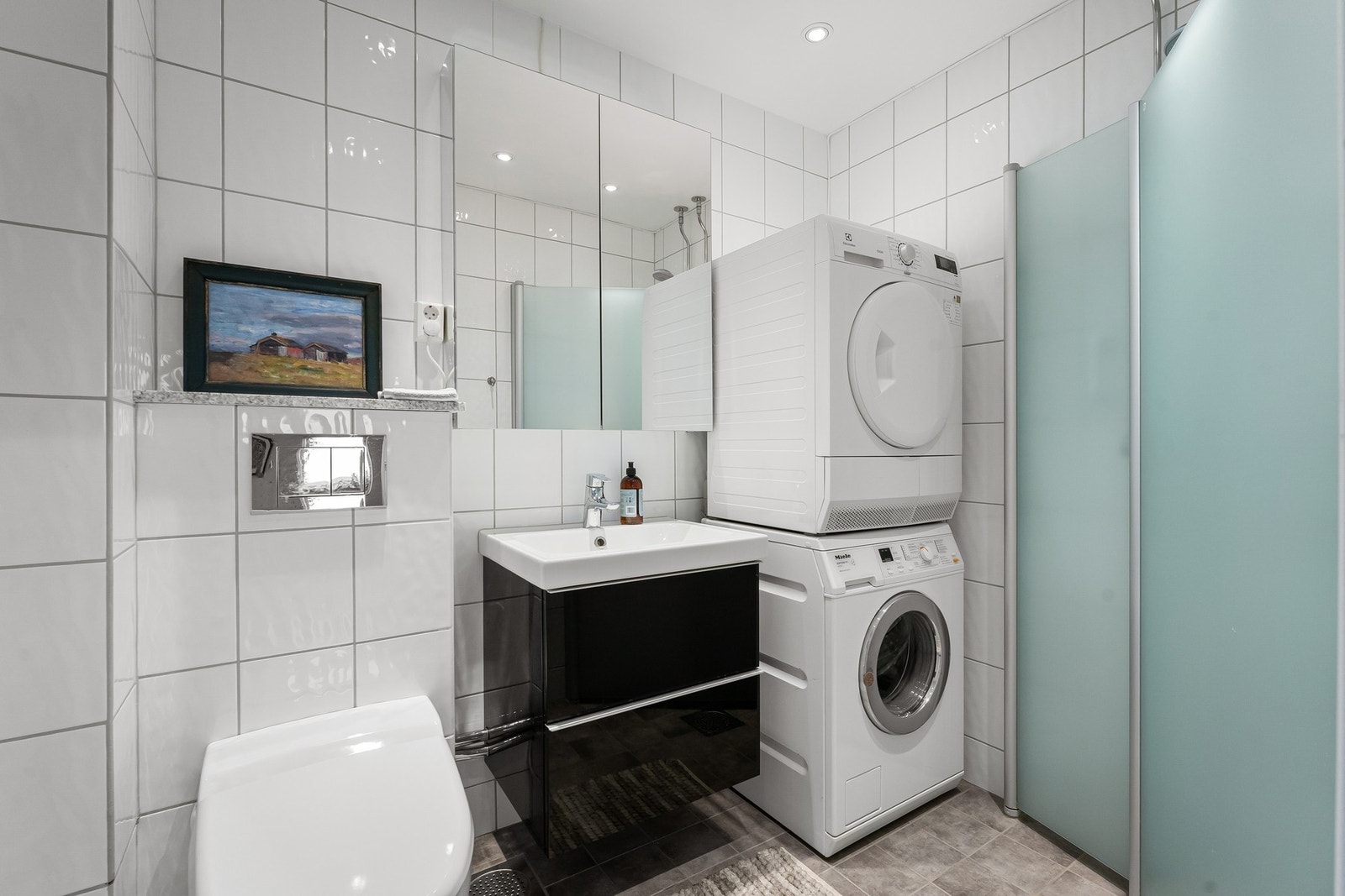 Badet fremstår som moderne og funksjonelt, med flislagte gulv og veggflater i lyse, tidløse fargetoner. Det er installert veggmontert toalett med innbyggingssisterne, servant med underskap og speilskap over servant. Galleribilde