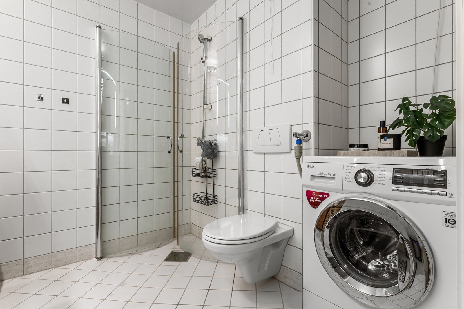 Badet er flislagt med varmekabler i gulv, veggmontert toalett og dusjhjørne med plassbesparende glassvegger som kan svinges inn utenom bruk. Opplegg til vaskemaskin og plass for tørketrommel i en praktisk nisje som gir maksimalt utbytte av plassen. Galleribilde