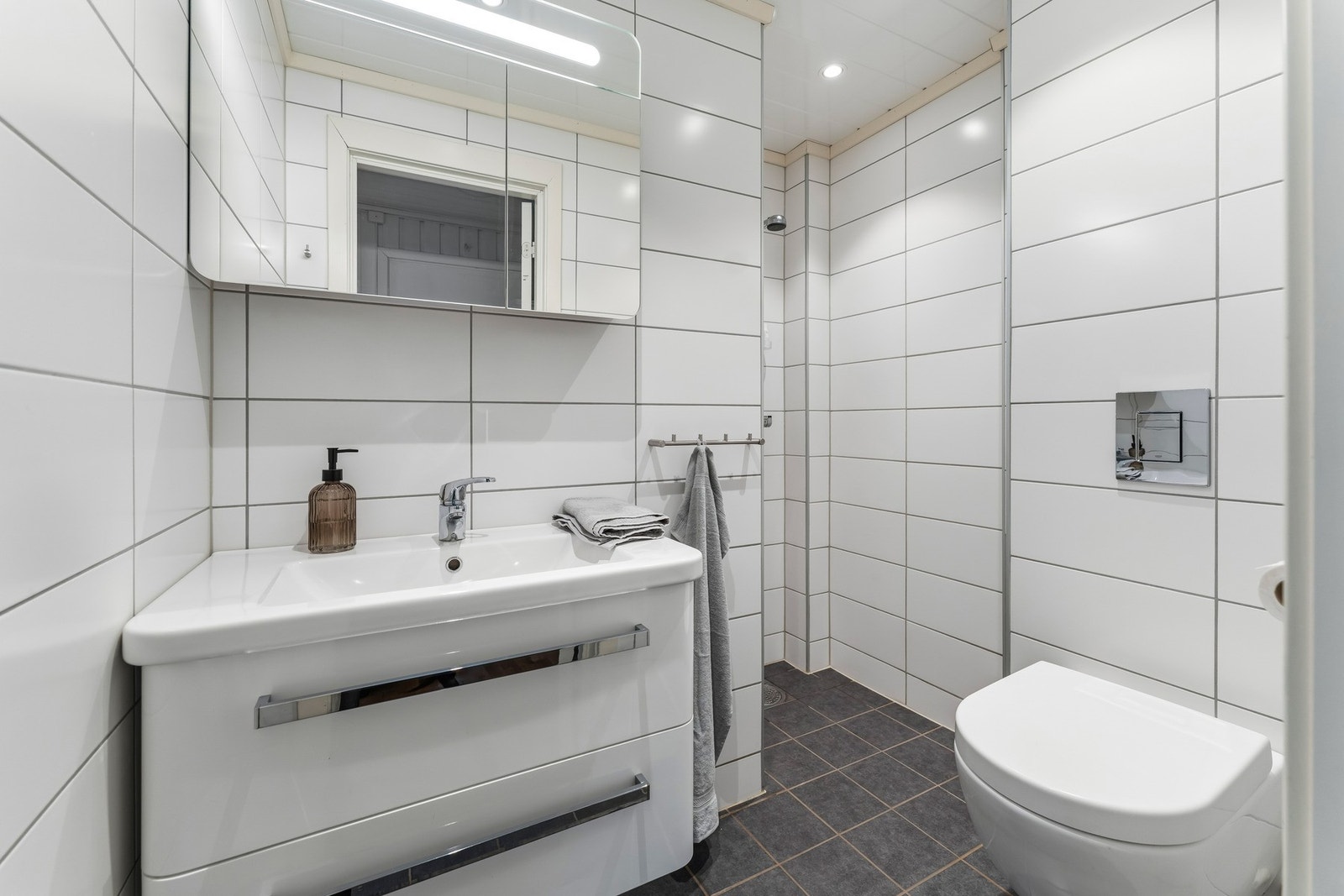 Flislagt bad med gulvvarme, pusset opp i 2017. Innredningen består av moderne servantinnredning med to skuffer, speilskap med lys og vegghengt toalett. Galleribilde