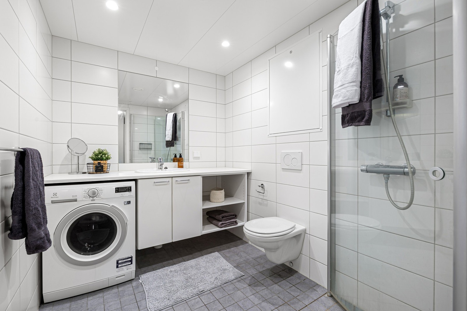 Leilighetens bad har flislagte overflater og elektriske varmekabler i gulv. Badet er utstyrt med innfellbare dusjdører i glass, vegghengt toalett, samt kran og avløp for vaskemaskin. Rørskapet er lokalisert over toalettet. Galleribilde