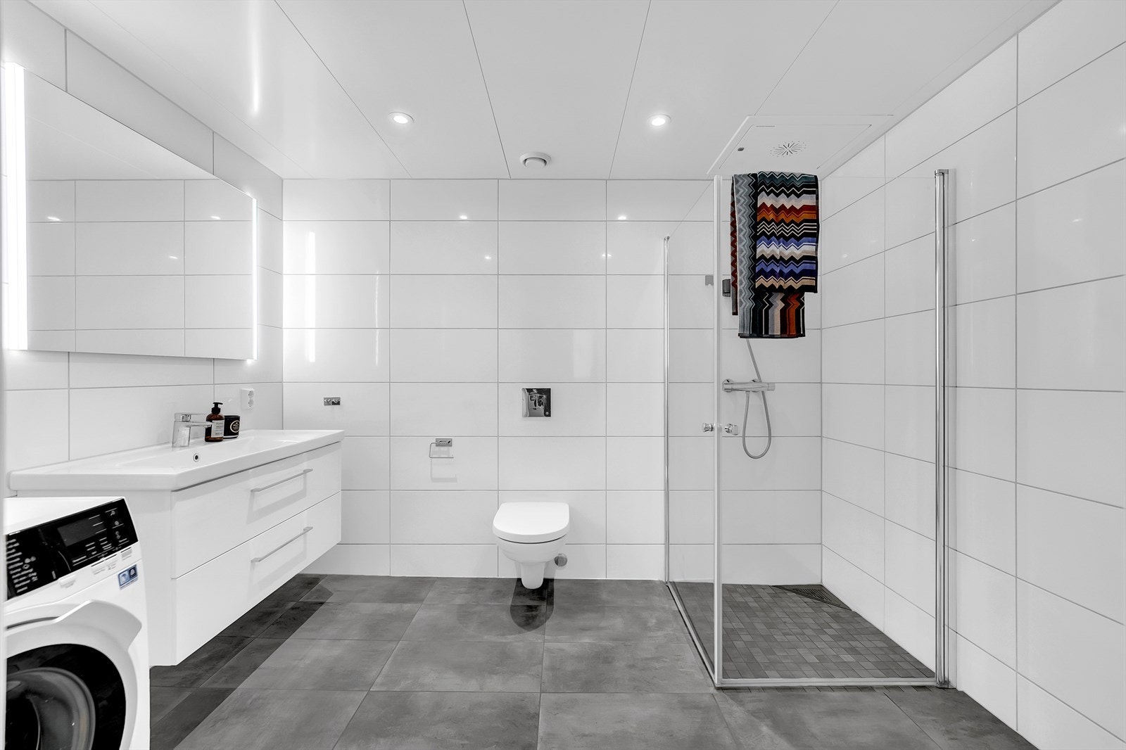 Flislagt baderom innredet med servant med underskap, dusjvegger, vegghengt toalett og opplegg for vaskemaskin. Galleribilde