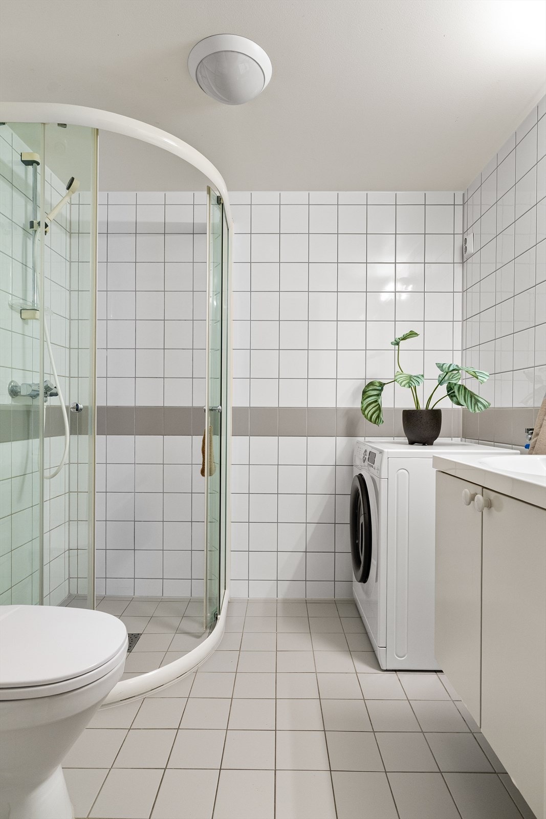 Badet er innredet med veggmontert dusjhjørne, servant med underskap, speil med integrert belysning, opplegg for vaskemaskin samt veggmontert wc. Galleribilde