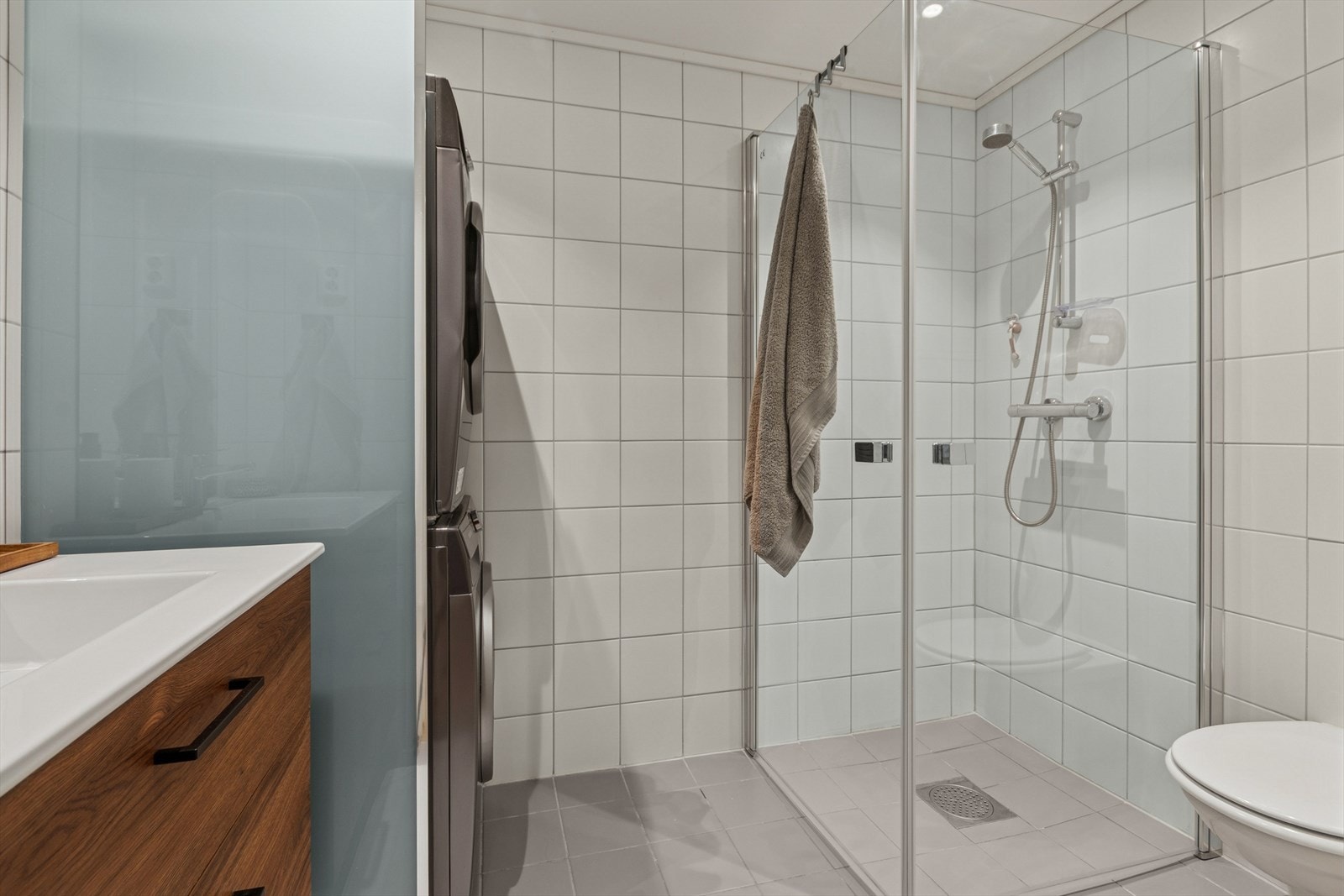 Badet er fra byggeår og har flislagte overflater på både vegger og gulv. Det er behagelig gulvvarme i rommet. Innredningen består av en servantseksjon med nedfelt vask, veggmontert toalett og dusjhjørne med dusjvegger Galleribilde