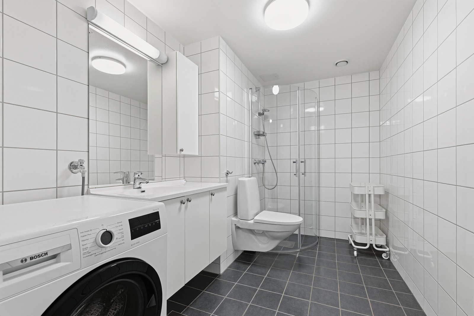 Pent flislagt bad med hjørnedusj, servant, toalett og vaskemaskin. Galleribilde