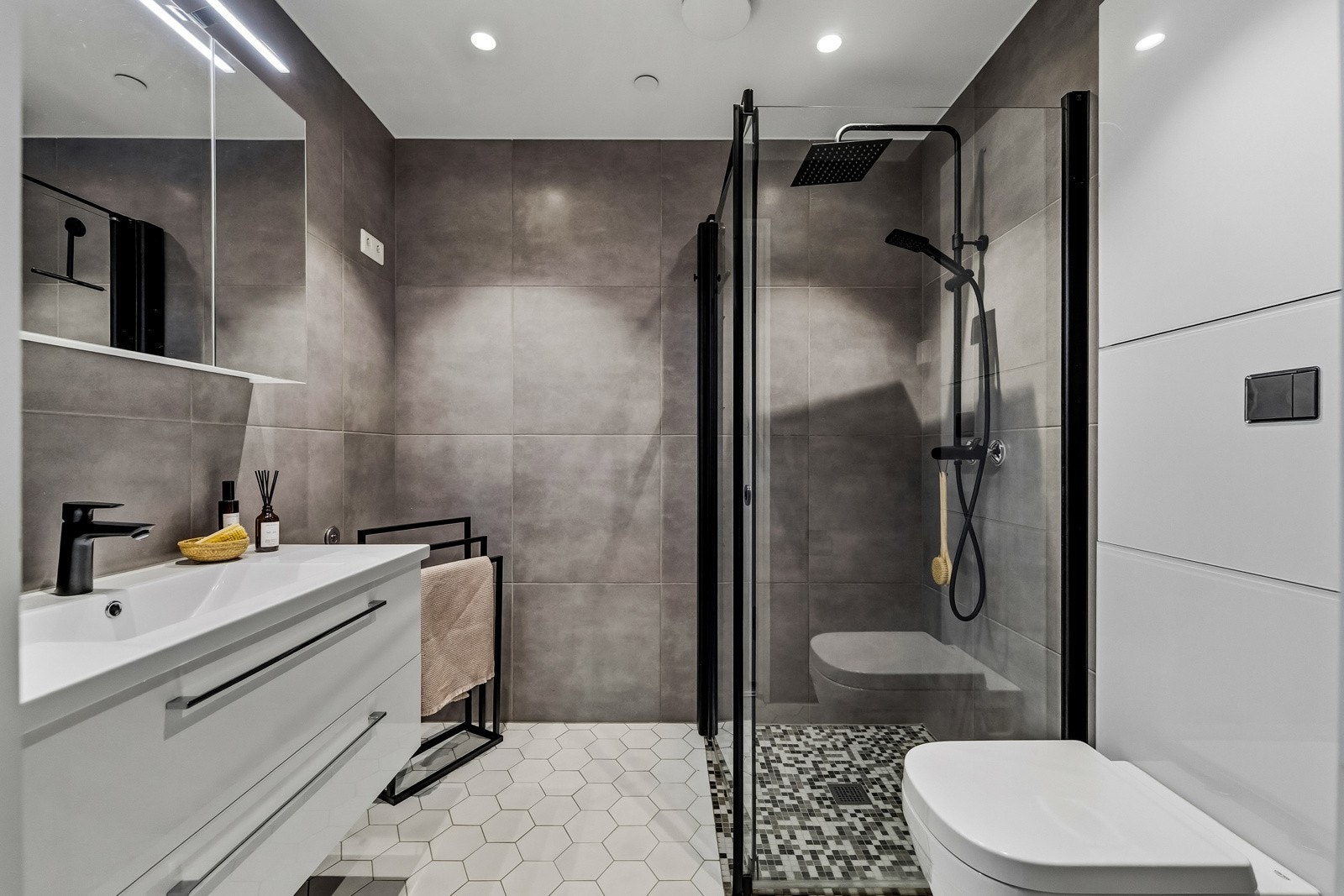 Badet er stilrent med flotte detaljer som sorte armaturer, mosaikkfliser i dusjen, grå veggfliser og hexagon gulvfliser. Det er gulvvarme (fjernvarme), downlights i himlingen, vegghengt toalett og et dusjhjørne med sorte detaljer Galleribilde