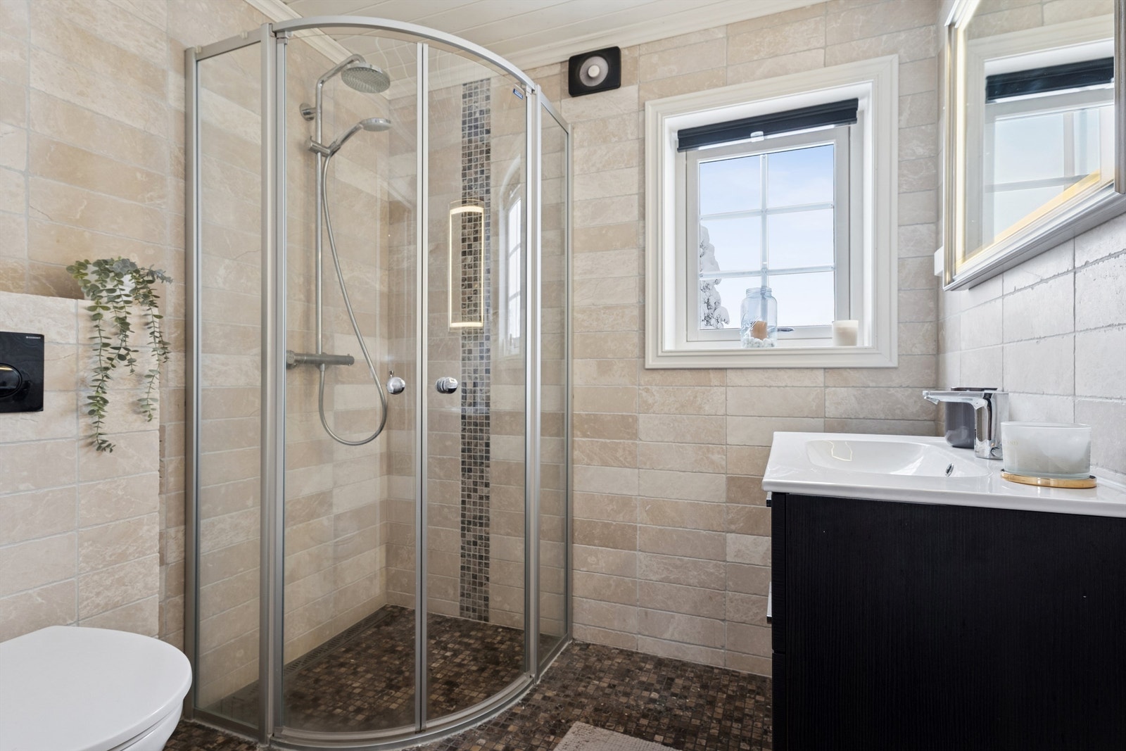 Badet er flislagt i stilrene farger og har gulvvarme, dusjhjørne, vegghengt toalett og servantinnredning. Galleribilde