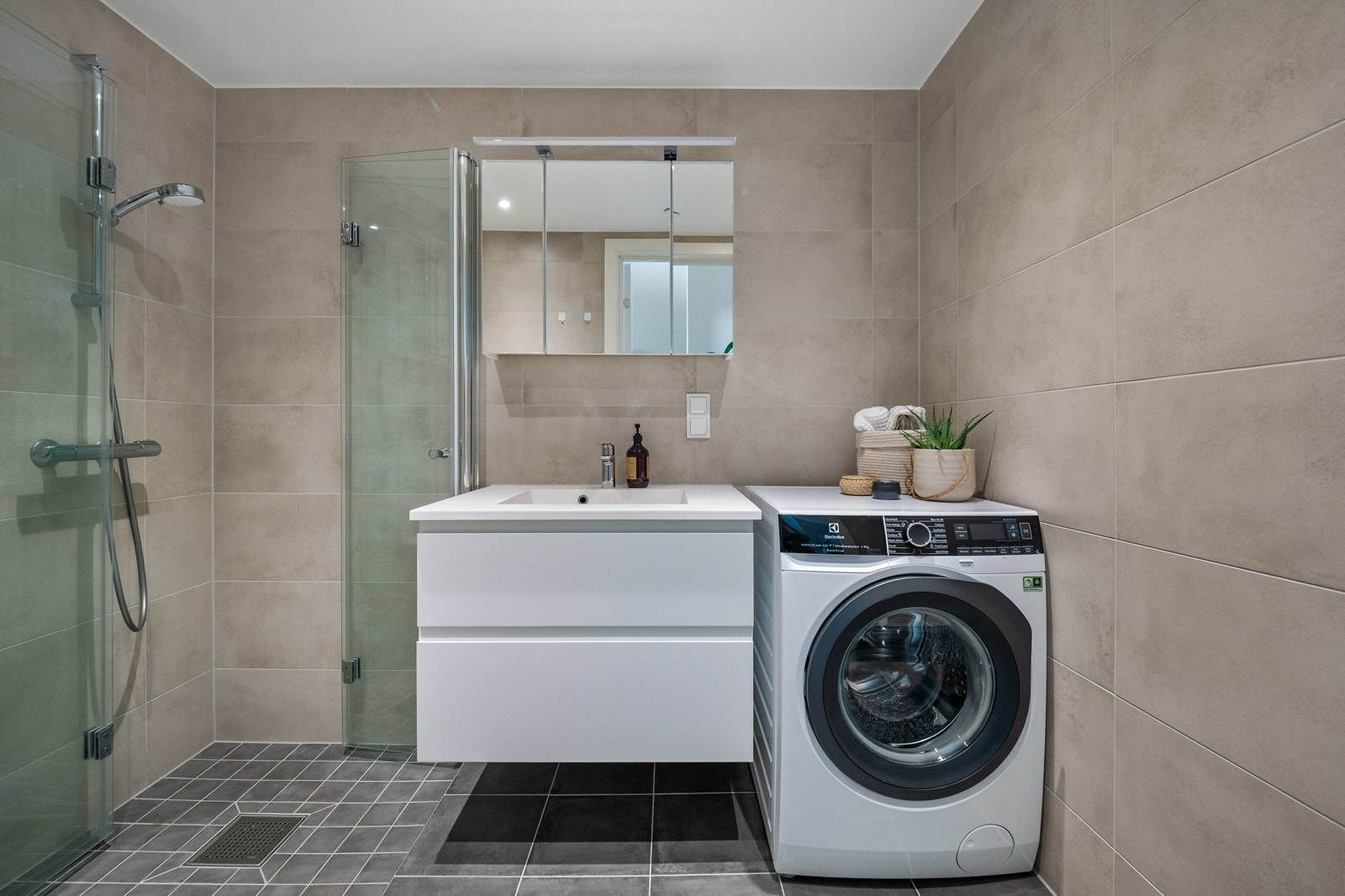 Flislagt bad med vegghengt servantinnredning, dusjnisje med glassdører, vegghengt toalett og opplegg for vaskemaskin Galleribilde