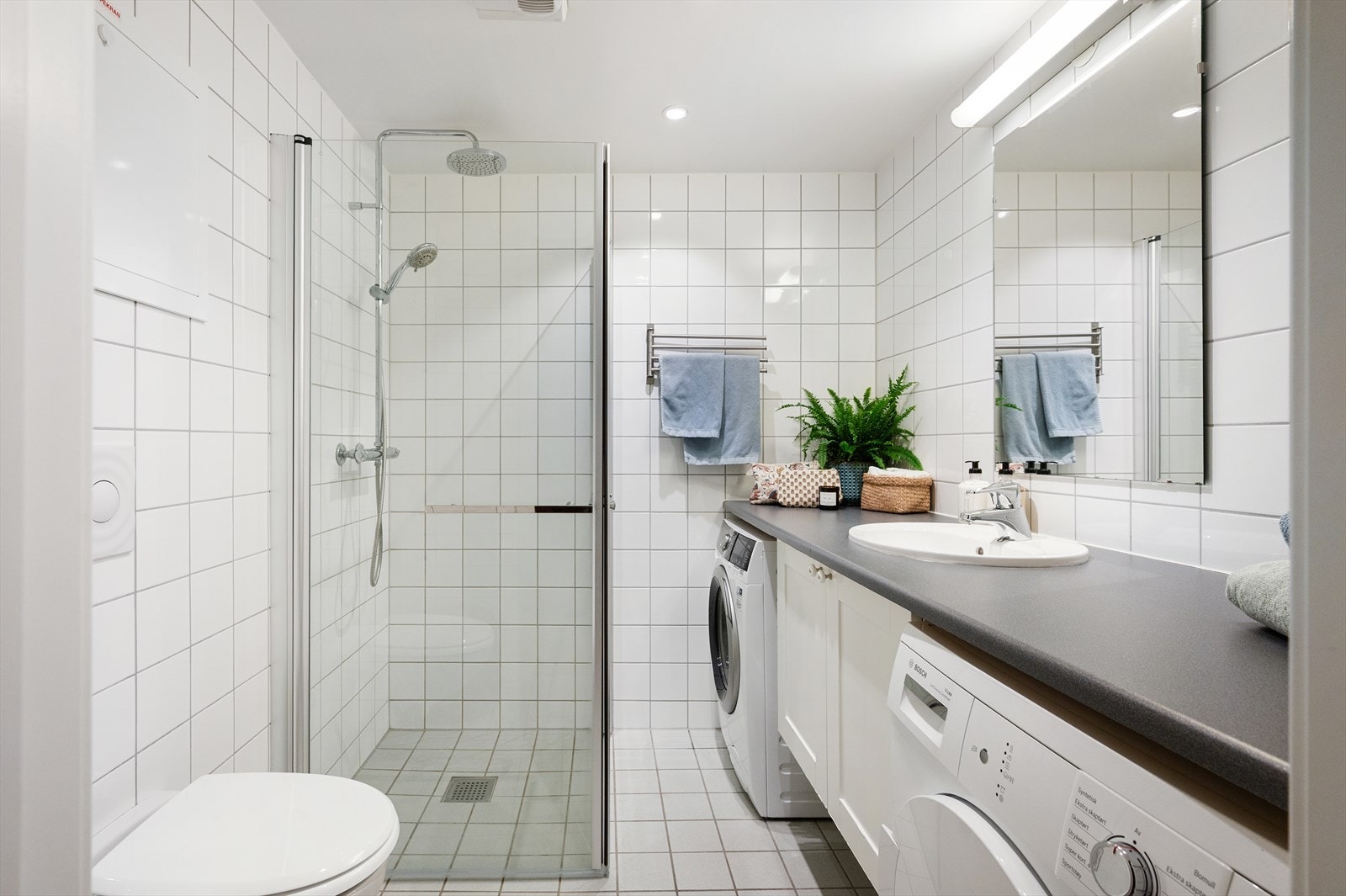 Pent og flislagt baderom innredet med vegghengt toalett, laminat benkeplate med nedfelt servant og underskap, samt dusjhjørne med svingbare glassdører. Galleribilde