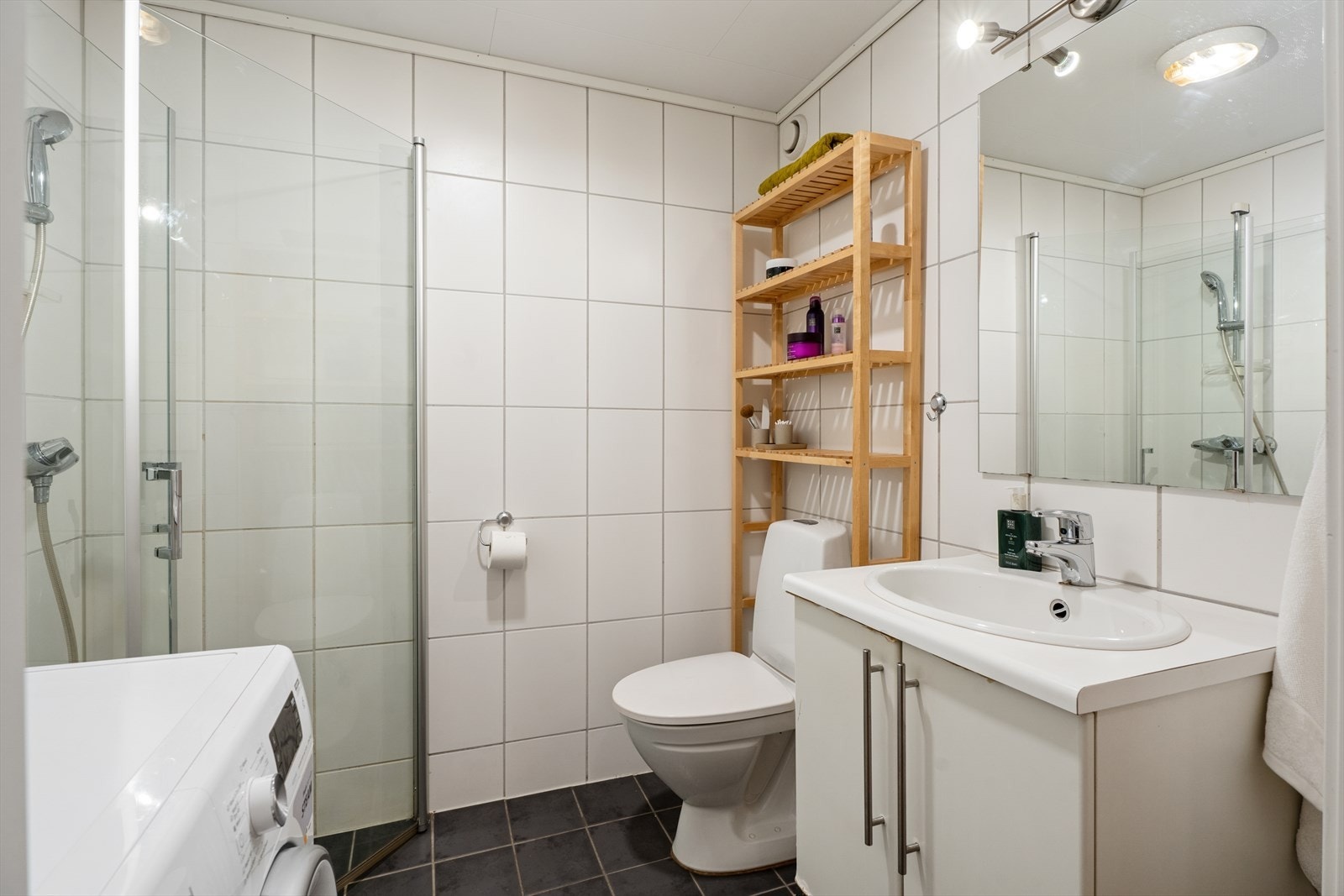 Baderom med flislagte overflater på gulv og vegger. Badet er innredet med baderomsmøbel med nedfelt servant med slette, lyse fronter og overhengende speil med belysning. Dusjen er plassert i hjørnet og har dører av glass og dusjarmatur med dusjsett. Galleribilde