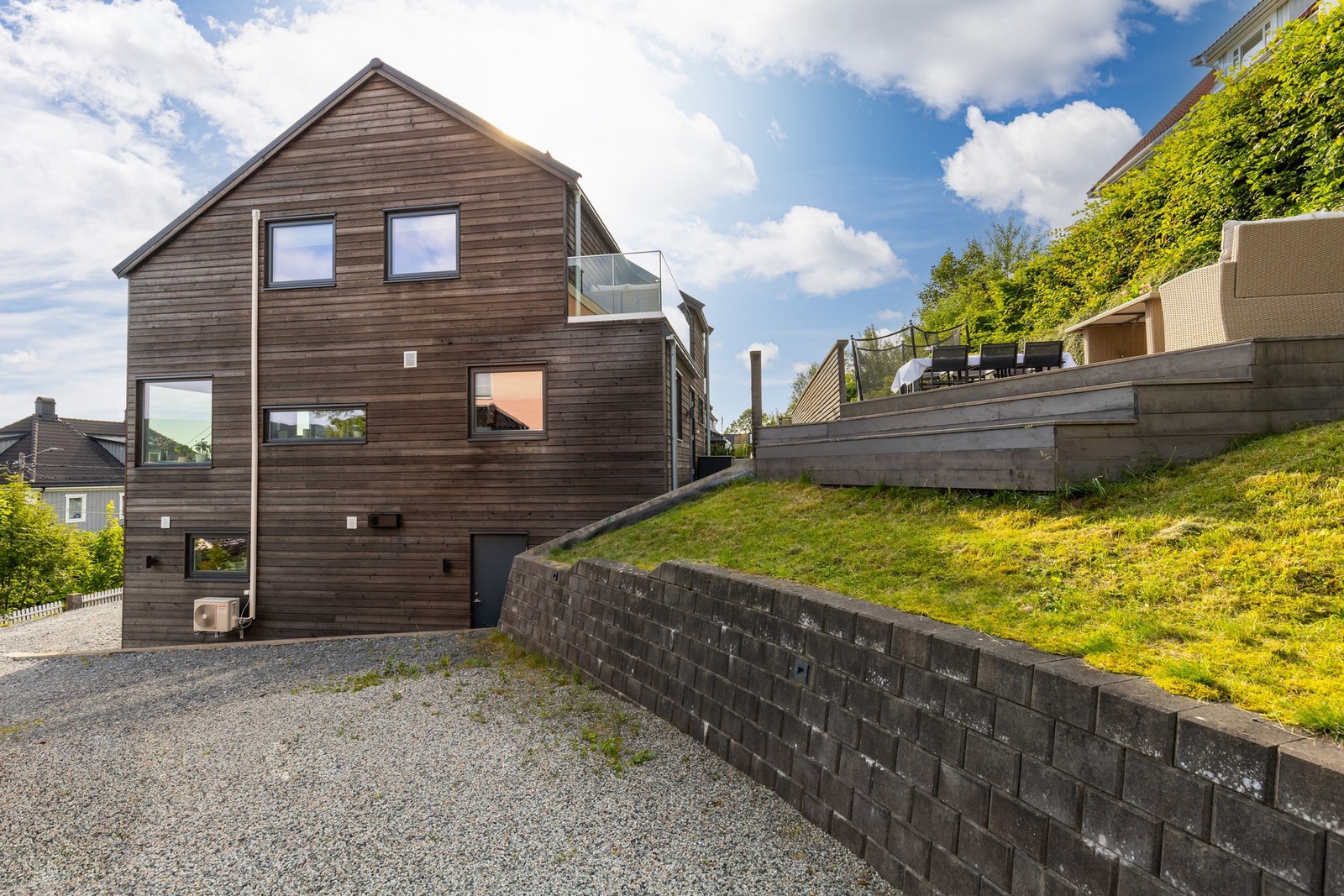 Boligen fremstår med et moderne og helhetlig uttrykk der trekledningen harmonerer med de solide støttemurene i forkant Galleribilde