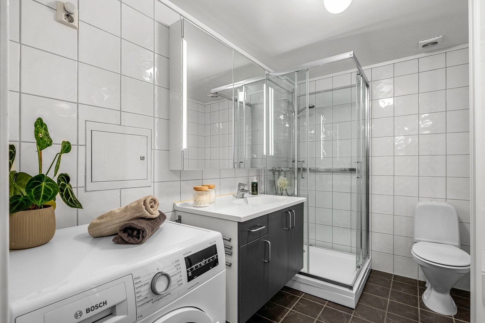 Badet er innredet med gulvmontert toalett, dusjkabinett, veggmontert servant og skapinnredning og speilskap, opplegg for vaskemaskin og fordelerskap til vannledninger Galleribilde