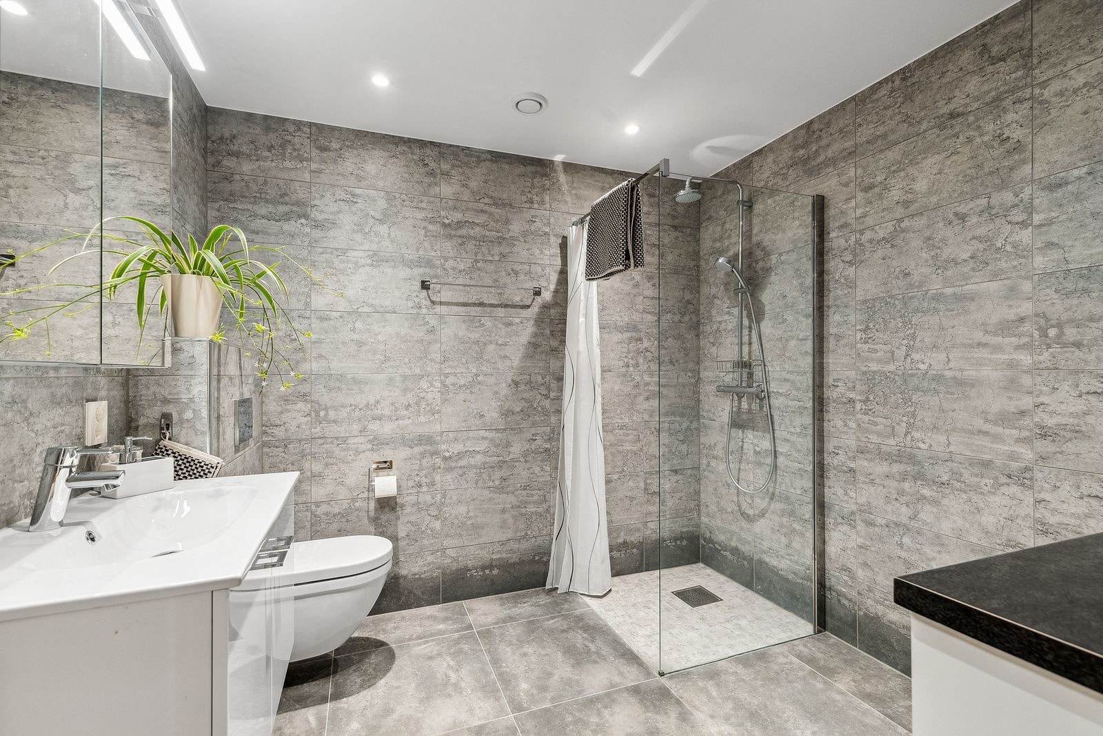 Flislagt bad i hybel med gulvvarme, downlights, vegghengt toalett, dusj med glassvegg, servant med underskap og opplegg for vaskemaskin. Galleribilde