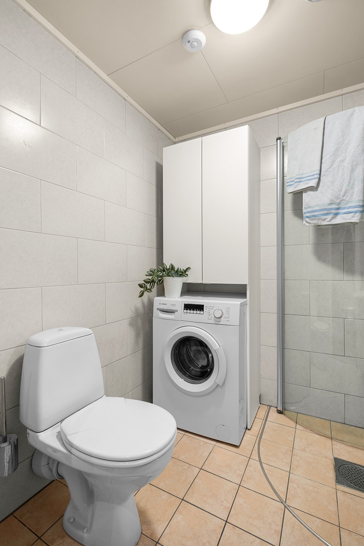 Badet er innredet med servantinnredning samt speil, gulvmontert toalett, dusjhjørne med dusjdører i glass og opplegg for vaskemaskin Galleribilde