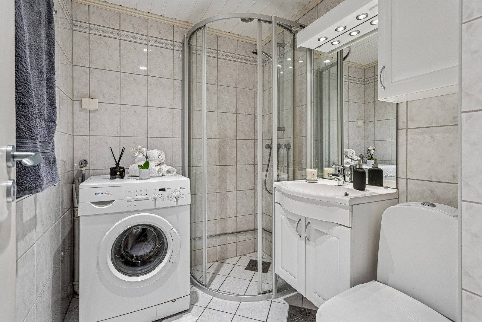 Badet er utstyrt med dusjhjørne med skyvedør, servant med underskap, speil med overlys, veggskap, toalett og opplegg for vaskemaskin. Galleribilde