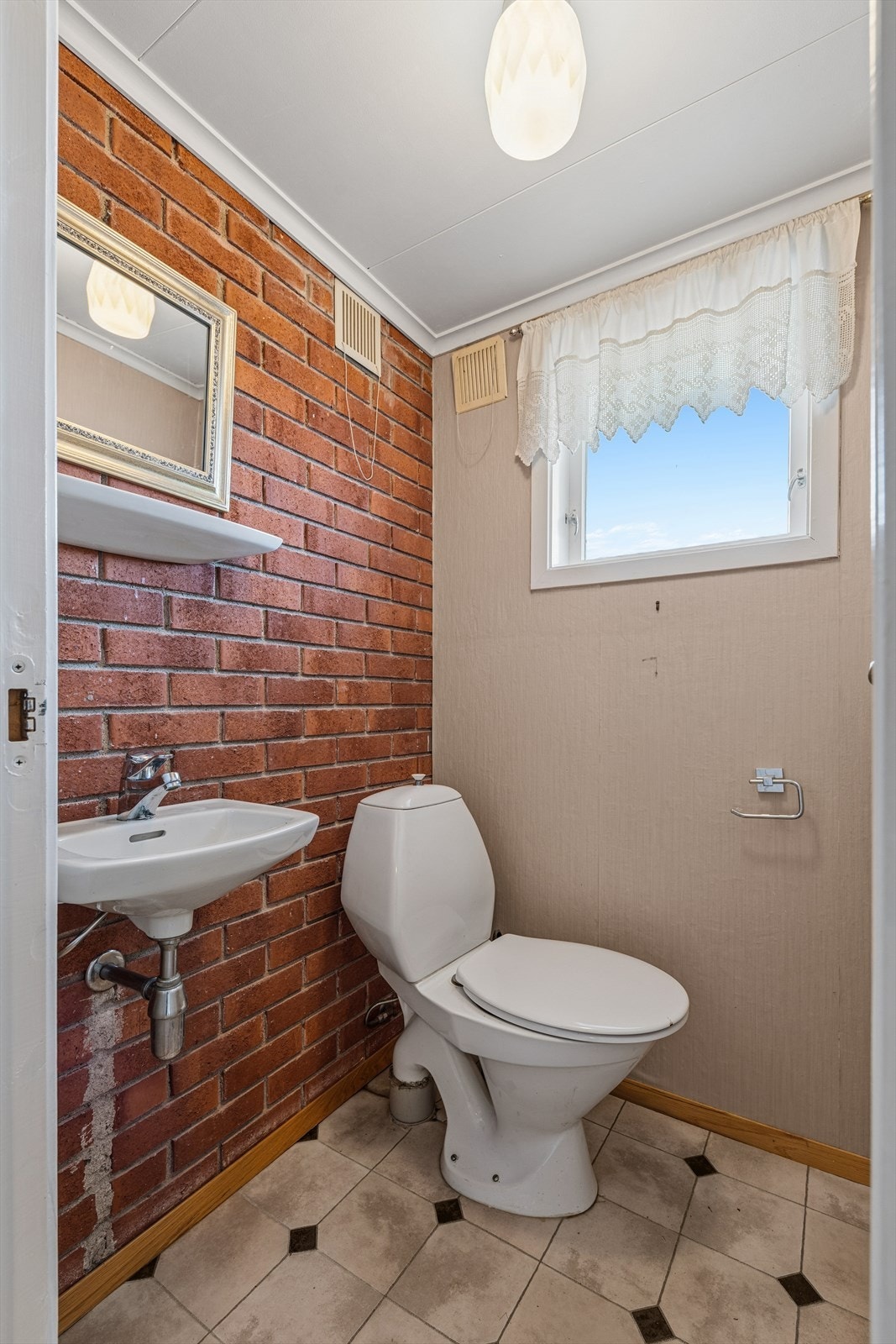 Toalettrom med belegg på gulv, malte tapetserte plater og teglstein på vegger, og malte takessplater i tak. Rommet er innredet med vegghengt servant og frittstående toalett. Galleribilde