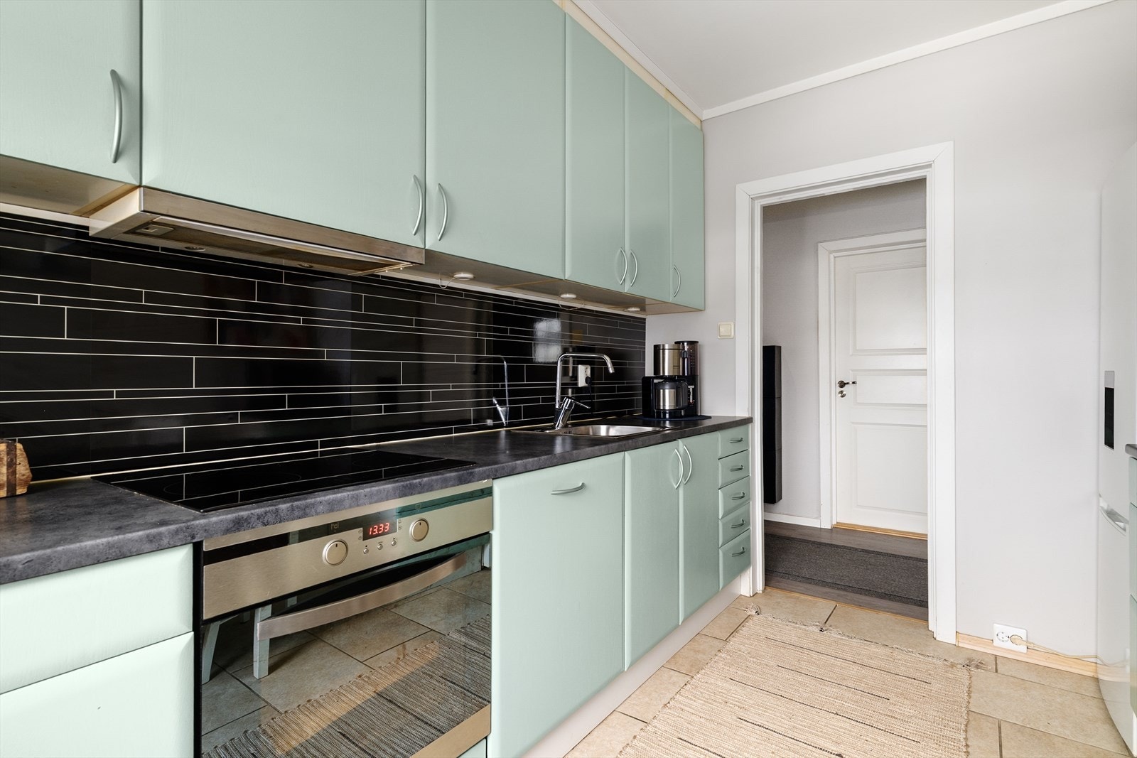 Beskyttelsesplater på vegg over laminat benkeplate med gode arbeidsflater, nedfelt platetopp og oppvaskkum i rustfritt stål. Avsatt plass til frittstående kjøleskap. Galleribilde