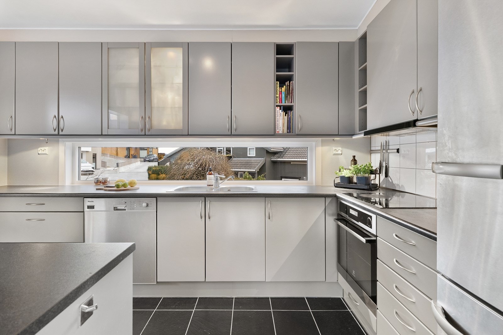 Innredningen har glatte, lysegrå fronter, frostet overskap, åpne hyller og laminert benkeplate med dobbel oppvaskkum. Over benken er det belysning, og over kokesonen har kjøkkenet hvite fliser og en ventilator med avtrekk ut. Galleribilde