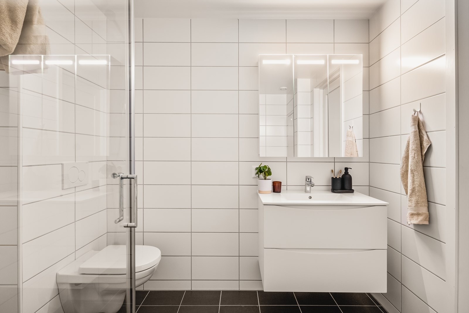 Badet har opplegg for vaskemaskin og tørketrommel, og er innredet med baderomsmøbel med heldekkende servant, speilskap m/ belysning, vegghengt toalett og dusj med glassdører. Galleribilde